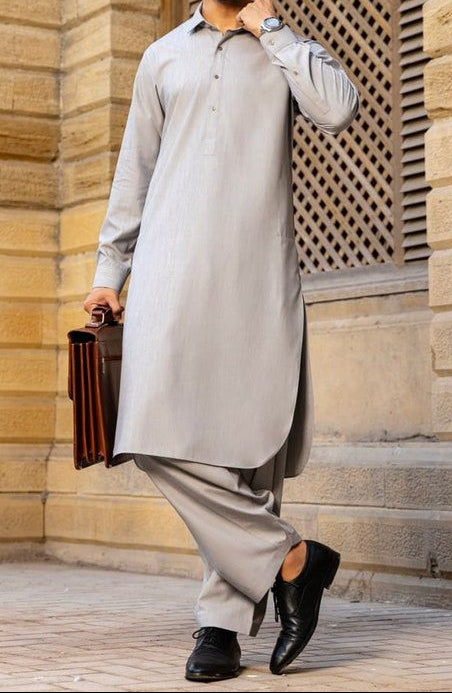 Man in a light gray traditional outfit standing in front of a stone building.