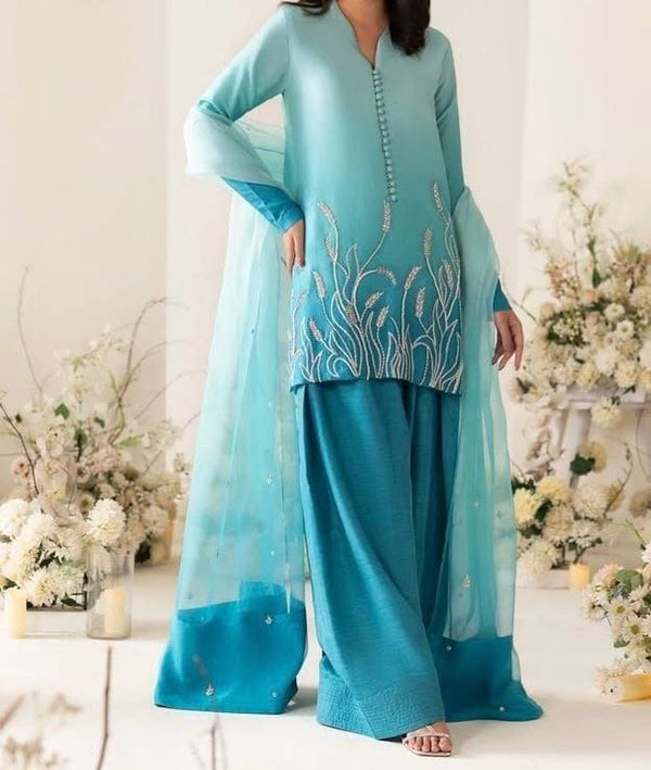 Woman wearing a turquoise traditional outfit with floral embroidery in a decorated indoor setting.