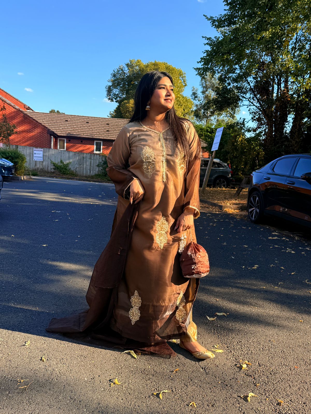 Woman in a long, patterned dress standing on a street with trees and houses in the background.
